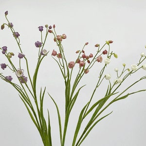 以下が含まれることがあります： 白い、ピンク、紫のベル型のつぼみを持つ、人工の花のブーケ。花は緑の茎に配置されています。