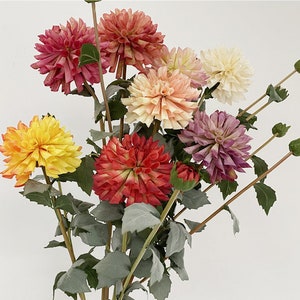 May include: A bouquet of artificial dahlias in various colors, including red, orange, yellow, pink, and purple. The flowers are arranged in a vase with green leaves.