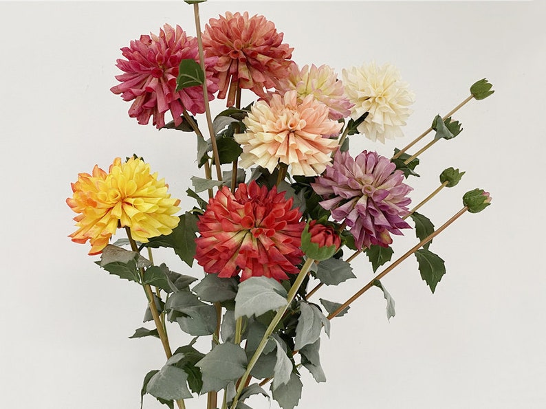 May include: A bouquet of artificial dahlias in various colors, including red, orange, yellow, pink, and purple. The flowers are arranged in a vase with green leaves.