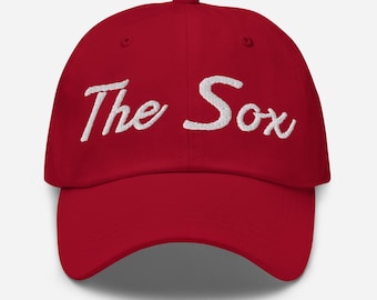 May include: A red baseball cap featuring the white embroidered text "The Sox". The cap has a curved peak and an adjustable fabric strap at the back.