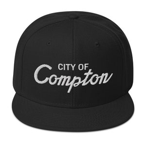May include: Black and white embroidered baseball cap with the text "CITY OF Compton" in a white cursive font.