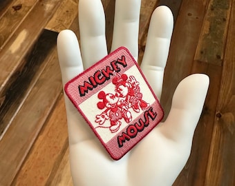 May include: A square, embroidered patch featuring Mickey and Minnie Mouse in red and white on a pink background. The words "Mickey Mouse" are embroidered in black. The patch is held in a white hand against a wooden background.