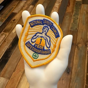 May include: A white and yellow embroidered patch with the text "National Safety Council" and "1 Year Safe Driver Award". The patch features a cartoon image of a person holding a yellow circle with the number "1" inside.