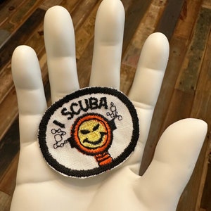 May include: A white hand holding a round, black and white embroidered patch with the word "SCUBA" and a smiling scuba diver with an orange tank and yellow face.