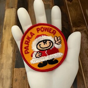 May include: A round, yellow and red embroidered patch with a cartoon character wearing a red and white parka and a peace sign. The text "PARKA POWER" is embroidered around the character.
