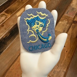 May include: A blue denim patch with an embroidered image of a yellow face blowing into a blue trumpet. The text "CHICAGO" is embroidered in blue below the image.