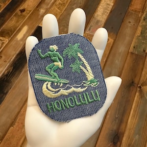 May include: A blue denim patch with green and white embroidery of a surfer riding a wave with palm trees in the background. The text "HONOLULU" is embroidered in green.