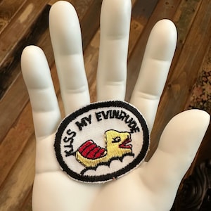 May include: A white hand holding a black and white embroidered patch with the text "Kiss My Evindun" and a yellow cartoon bird with red and white stripes.