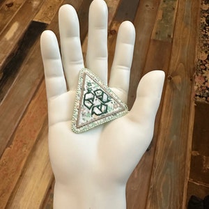 May include: A triangular green and white embroidered patch with a recycling symbol in the center. The patch is on a white hand.