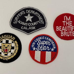 May include: Four embroidered patches. A black patch with gold embroidery reads "JAGUAR" with a stylized jaguar head and checkered pattern. A blue patch with silver embroidery reads "SURFER'S DEPARTMENT ADAMS COUNTY CAL.ASK" with a silver star. A red patch with white embroidery reads "I'M THE BEAUTIFUL BRUTE". A blue, red, and white patch with white embroidery reads "1991 COOKIE CAMPAIGN Super Star" with a star and red and white stripes.