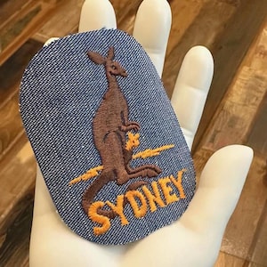 May include: A blue denim patch with an embroidered brown kangaroo and the word "SYDNEY" in orange thread.