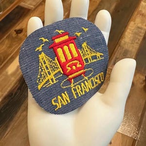 May include: A blue denim patch with an embroidered image of a red cable car and the Golden Gate Bridge. The text "SAN FRANCISCO" is embroidered in yellow below the image.