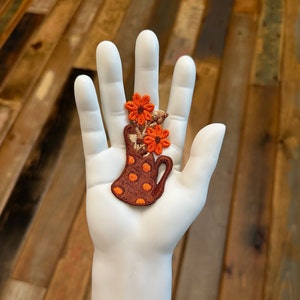 May include: A brown and orange embroidered patch of a pitcher with orange flowers. The pitcher has white polka dots.