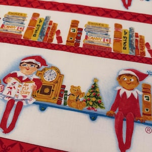 May include: White fabric with a repeating Christmas-themed pattern. The design includes elves, clocks, books labeled "Cookies," "Candy," and "Dolls," and a Christmas tree. Red decorative borders frame the design. The text "AN ELF STORY" is visible.