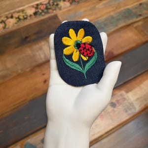 May include: Embroidered patch featuring a yellow flower and a red ladybug on a dark blue background. The patch is oval-shaped and has green leaves and stem. The patch is held by a white hand.