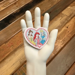 May include: A heart-shaped patch with a pink border and white polka dots features three Disney princesses: Belle, Ariel, and Cinderella. The princesses are wearing their iconic dresses and are standing together.