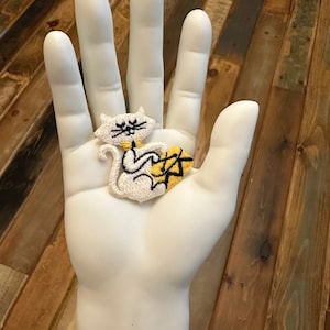 May include: White embroidered iron-on patch of a cat holding a ball of yarn. The cat is white with black eyes and a black nose. The yarn is yellow and black.