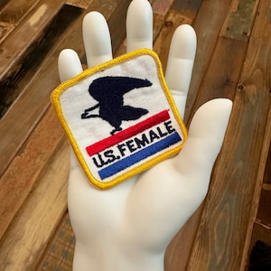 May include: A white fabric patch with a blue eagle and red, white, and blue stripes. The text "U.S. FEMALE" is embroidered in blue on the patch.