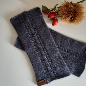 Könnte beinhalten: Ein Paar graue gestrickte Fingerlose Handschuhe mit einem Zopfmuster und braunen Leder-Etiketten mit der Aufschrift "Handgefertigt".