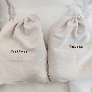 May include: Two beige drawstring produce bags, one labeled "Potatoes" and the other "Onions" in black text. The bags are made of a natural fabric and are on a marble surface. A wooden crate of produce is visible on the left.