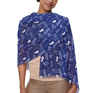 May include: A blue scarf with a white pattern of horseshoes, cowboy hats, and the word "BLUE" repeated throughout. The scarf is draped around the shoulders of a person wearing a tan top and pants.