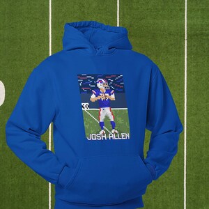 K&ouml;nnte beinhalten: Ein k&ouml;nigsblauer Hoodie mit einem pixeligen Bild von Josh Allen, einem Footballspieler in einem blau-roten Trikot, auf der Vorderseite. Das Bild ist vor einem blau-wei&szlig;en Hintergrund mit Konfetti platziert. Der Text "Josh Allen" ist unter dem Bild gedruckt.