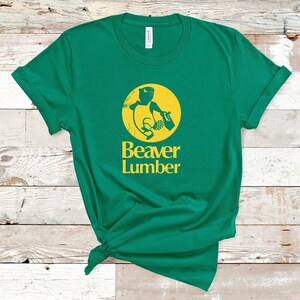 Op de afbeelding: Een groen T-shirt met een geel logo van een bever die een boomstam vasthoudt. Het logo bevindt zich in een cirkel met de tekst "Beaver Lumber" eronder.