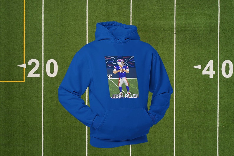 K&ouml;nnte beinhalten: Ein k&ouml;nigsblauer Hoodie mit einem pixeligen Bild von Josh Allen, einem Footballspieler in einem blau-roten Trikot, auf der Vorderseite. Das Bild ist vor einem blau-wei&szlig;en Hintergrund mit Konfetti platziert. Der Text "Josh Allen" ist unter dem Bild gedruckt.