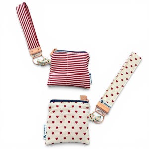 May include: Three small fabric pouches with wrist straps. One pouch is white with red hearts, another is red and white striped, and the third is red and white striped. Each pouch has a zipper and a small tag that says "Alkmini's".