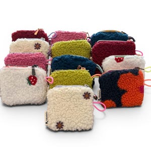 May include: A collection of small, fluffy, zippered pouches in various colors, including cream, pink, green, and navy. Some pouches feature embroidered floral or fruit designs. Each pouch has a small loop and a zipper.