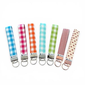 Könnte beinhalten: Sieben Schlüsselanhänger mit Stoffbändern in verschiedenen Mustern, darunter Vichy, Streifen und Herzen. Die Farben sind Blau, Rosa, Orange, Grün und Rot. Jeder Schlüsselanhänger hat einen Metallring und den Text "Alkmini's" auf dem Band.