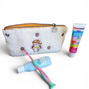 Könnte beinhalten: Eine weiße, flauschige Kosmetiktasche mit einem gestickten Hasen- und Blumendesign, eine rosa-grüne Zahnbürste, eine blaue Flasche und eine Tube Kinderzahnpasta mit Peppa Wutz-Figuren.