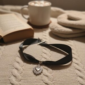 Può includere: Una collana girocollo in nastro nero con un ciondolo a cuore argentato. La collana è esposta su un maglione a trecce color crema. Una tazza di cioccolata calda, un libro e dei guanti sono sullo sfondo.