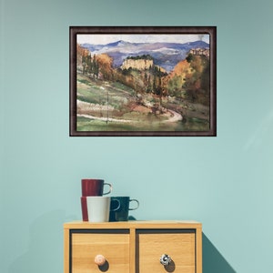 K&ouml;nnte beinhalten: Ein gerahmtes Aquarellbild, das eine Landschaft mit sanften H&uuml;geln, B&auml;umen und einer fernen Bergkette zeigt. Das Bild ist in einem braunen Rahmen und h&auml;ngt an einer hellblauen Wand.