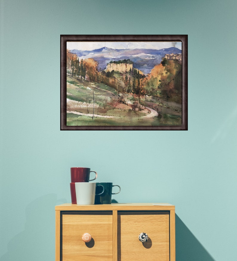 K&ouml;nnte beinhalten: Ein gerahmtes Aquarellbild, das eine Landschaft mit sanften H&uuml;geln, B&auml;umen und einer fernen Bergkette zeigt. Das Bild ist in einem braunen Rahmen und h&auml;ngt an einer hellblauen Wand.