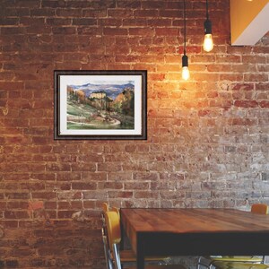K&ouml;nnte beinhalten: Ein gerahmtes Aquarellbild einer Landschaft mit einem kleinen Dorf in der Ferne. Das Bild ist vor einer Backsteinwand mit einem Holztisch und gelben St&uuml;hlen im Vordergrund platziert.