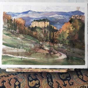 K&ouml;nnte beinhalten: Aquarellmalerei einer Landschaft mit einer Felswand, B&auml;umen und einer kurvigen Stra&szlig;e. Das Gem&auml;lde ist in Gr&uuml;nt&ouml;nen, Braun- und Blaut&ouml;nen gehalten. Die Unterschrift des K&uuml;nstlers ist unten rechts sichtbar.
