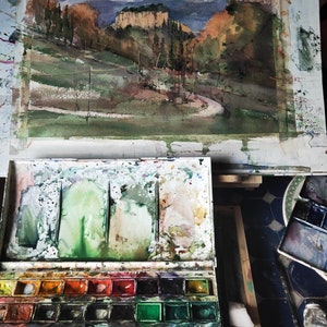 K&ouml;nnte beinhalten: Ein Aquarellbild einer Landschaft mit einer kurvigen Stra&szlig;e, die zu einer Felswand f&uuml;hrt. Das Bild steht auf einer Staffelei und im Vordergrund befindet sich eine Aquarellpalette mit Farbt&ouml;pfen und Pinseln.