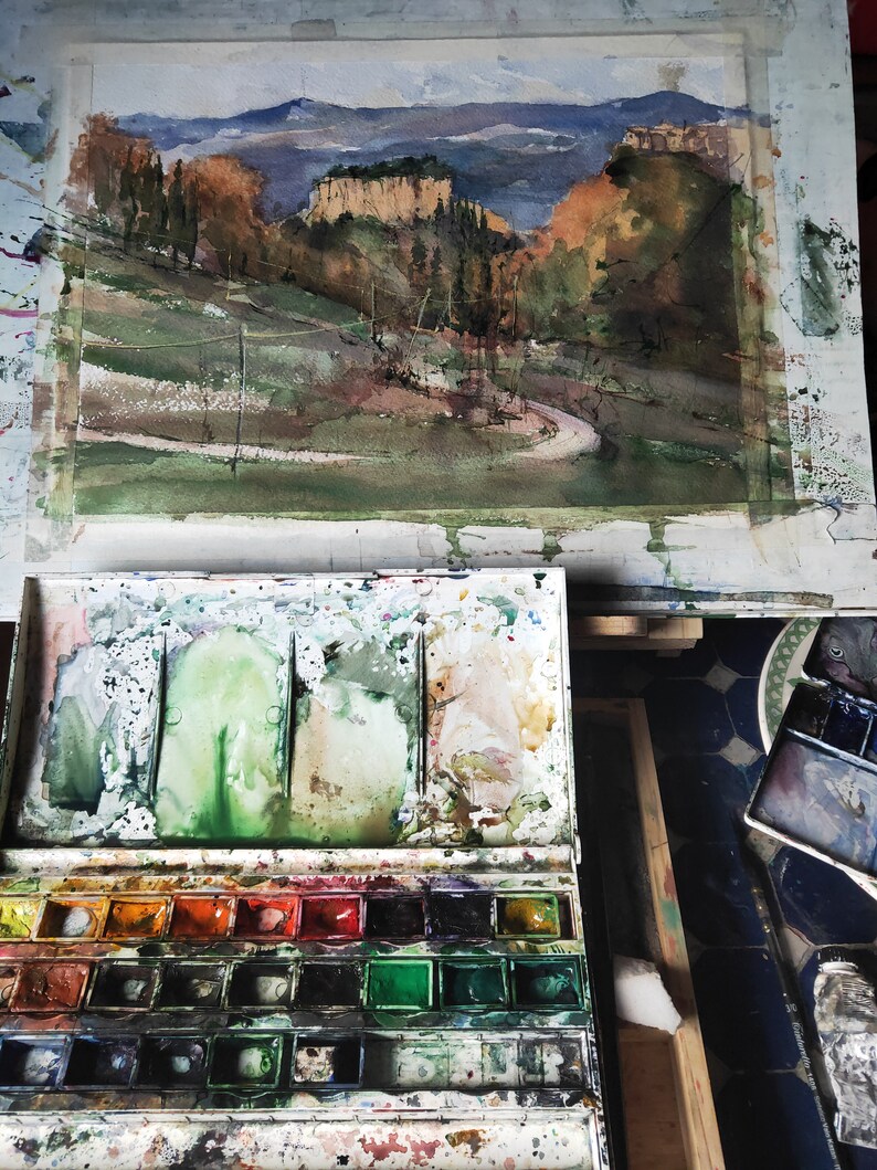 K&ouml;nnte beinhalten: Ein Aquarellbild einer Landschaft mit einer kurvigen Stra&szlig;e, die zu einer Felswand f&uuml;hrt. Das Bild steht auf einer Staffelei und im Vordergrund befindet sich eine Aquarellpalette mit Farbt&ouml;pfen und Pinseln.