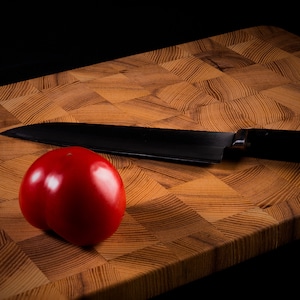 Puede incluir: Un tomate rojo y un cuchillo de chef negro sobre una tabla de cortar de madera.