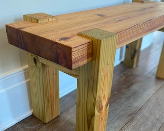 May include: A rustic wooden bench with a natural wood finish. The bench has a rectangular top and four legs. The legs are made of thick, square pieces of wood. The bench is made of reclaimed wood.