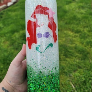 Op de afbeelding: Een witte tumbler met een groene glitterbodem. De bovenste helft van de tumbler heeft een roodharige zeemeermin met een paarse schelpbeha en een roze bloem in haar haar.