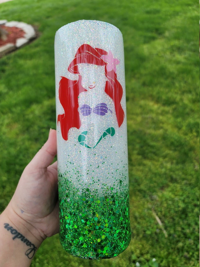 Op de afbeelding: Een witte tumbler met een groene glitterbodem. De bovenste helft van de tumbler heeft een roodharige zeemeermin met een paarse schelpbeha en een roze bloem in haar haar.