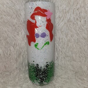 Op de afbeelding: Een witte tumbler met een groene glitterbodem en een witte glittertop. De tumbler heeft een rode, paarse en groene silhouet van Ariel, de Kleine Zeemeermin, aan de voorkant.