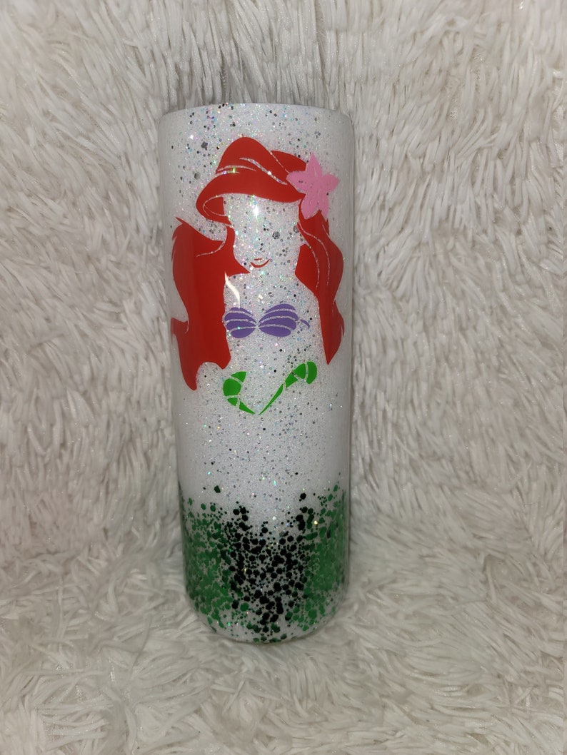 Op de afbeelding: Een witte tumbler met een groene glitterbodem en een witte glittertop. De tumbler heeft een rode, paarse en groene silhouet van Ariel, de Kleine Zeemeermin, aan de voorkant.