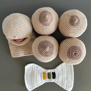 Op de afbeelding: Een verzameling handgemaakte haakartikelen, waaronder borstvormige vormen in verschillende tinten beige en bruin, een gezicht en een witte vlinderdas met gekleurde strepen. De items zijn gerangschikt op een grijze ondergrond.