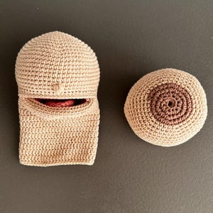 May include: A beige crochet object shaped like a hooded garment with a small opening revealing a red interior, positioned next to a round, beige crochet sphere featuring a darker brown circular center that resembles a human breast anatomy.
