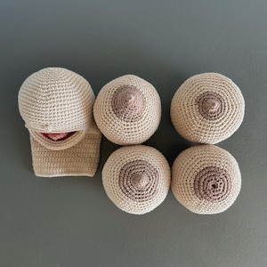 May include: A collection of handmade, beige crochet breast forms and a matching crochet head and neck piece. The breasts have a darker brown areola and nipple detail. The head and neck piece has a pink interior.