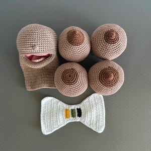 May include: A collection of handmade crochet items, including a beige face mask with a pink interior, four breast-shaped objects, and a white bow tie with yellow, green, and black stripes. The items are arranged on a gray surface.