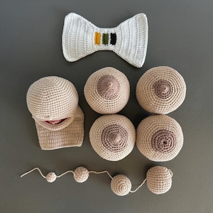 May include: Handmade crochet items including a white bow tie with green, yellow, and black stripes, several beige breasts, and a string of beige crochet balls. The items are arranged on a gray surface.